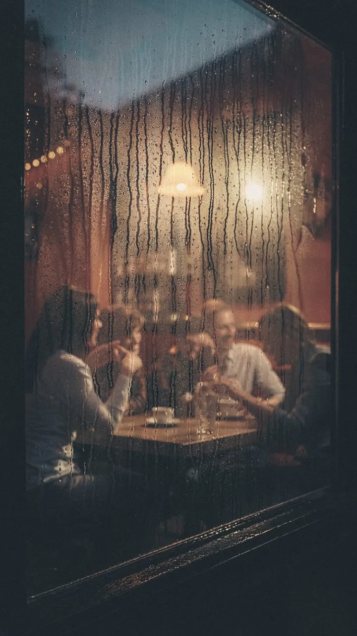A warm room at night seen through a rain-streaked window, people talking round a table.