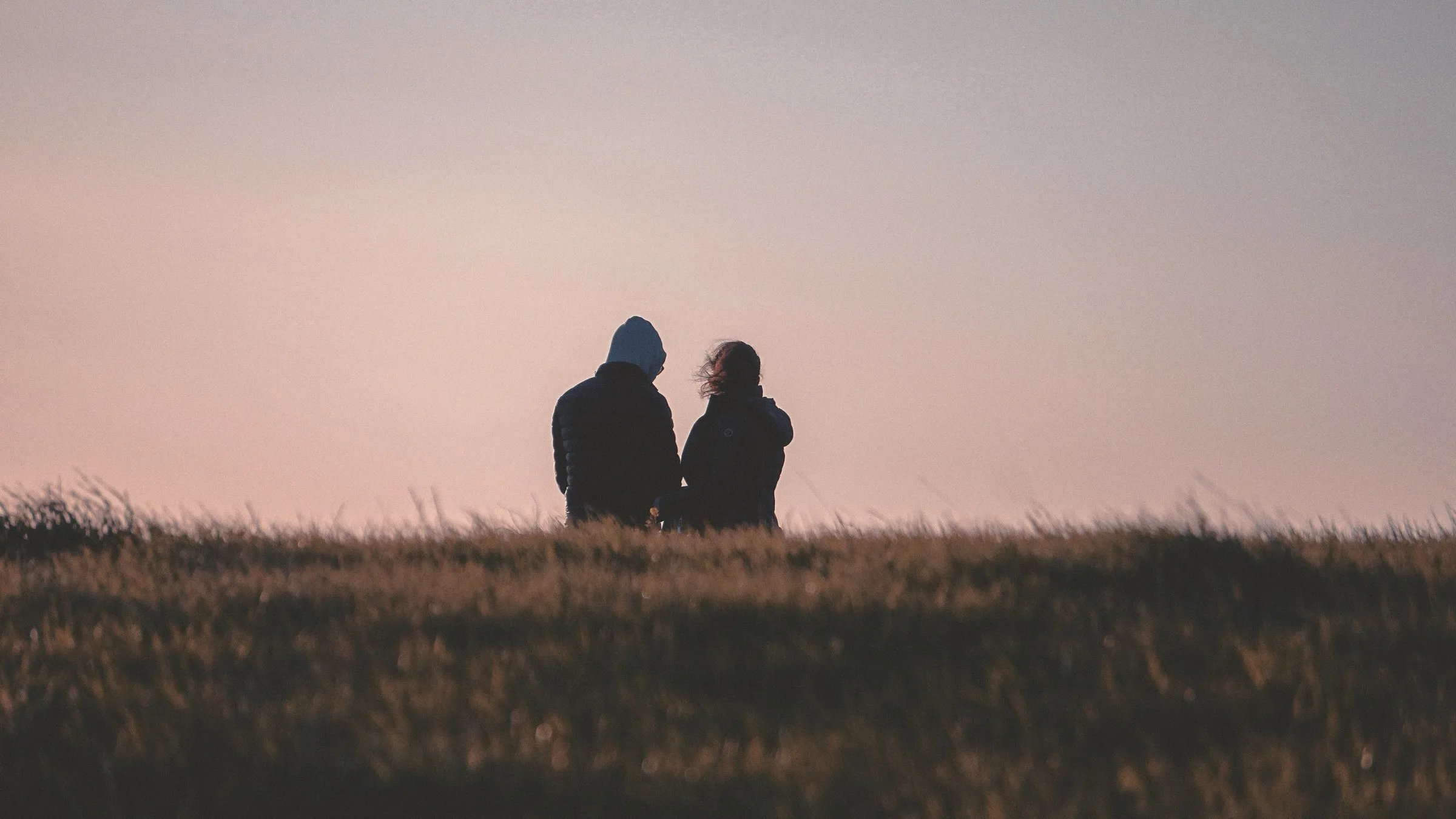 Two people sitting close on a grassy hill under a pink dusk sky.