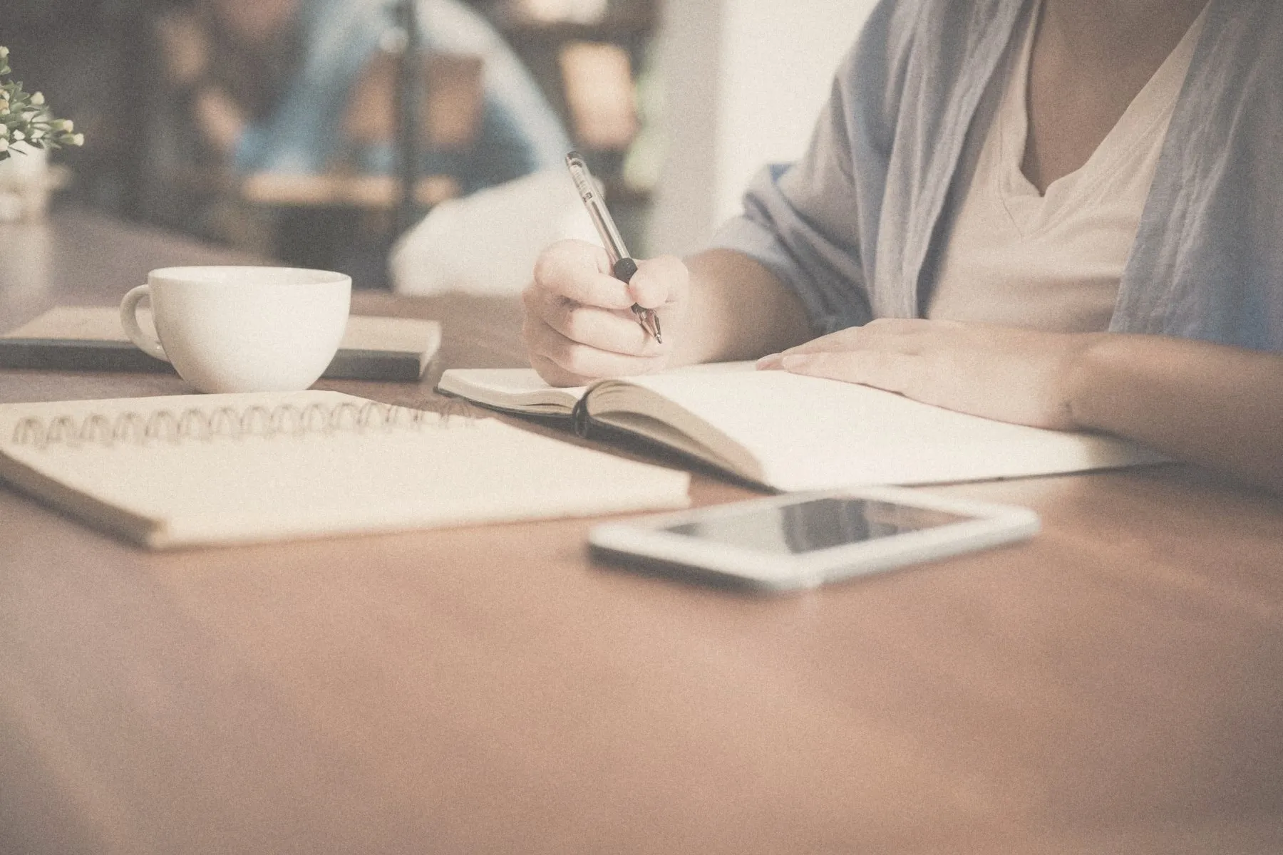 Someone writing in a notebook at a table with a cup of coffee