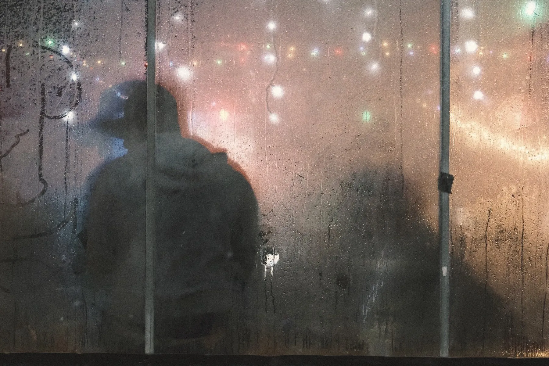 A figure behind a steamed-up café window, fairy lights blurred beyond